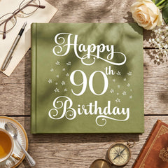 a green book titled "Happy 90th Birthday" with white text, placed on a wooden surface alongside other items such as a rose, a compass, and a teacup.