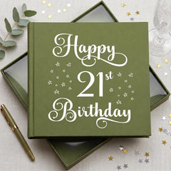 A green book titled "Happy 21st Birthday" with white text and stars on the cover, placed on a table with a pen, a glass, and some gold and silver confetti.