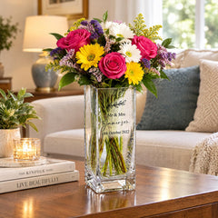 A clear glass vase filled with a colorful bouquet of flowers, including roses, daisies, and sunflowers, sits on a wooden table in a cozy living room.