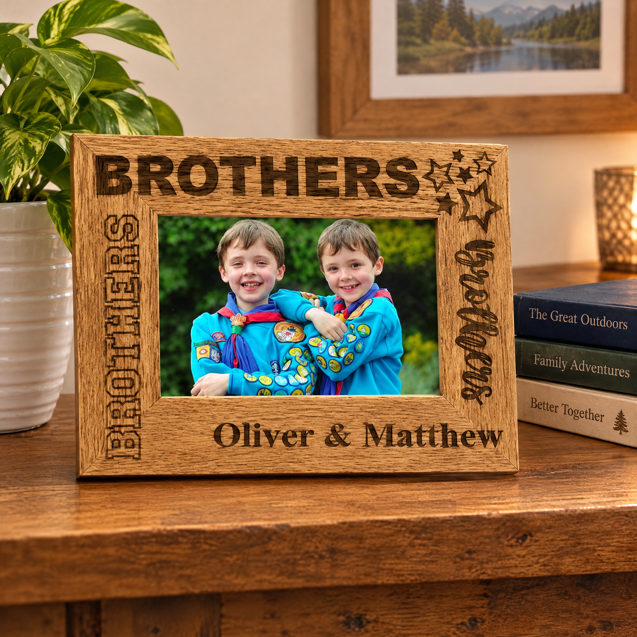 A wooden picture frame with the names "Brothers" and "Oliver & Matthew" engraved on it, holding a photo of two smiling boys. The frame is placed on a wooden surface, with a potted plant and a stack of books visible in the background.