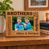 A wooden picture frame with the names "Brothers" and "Oliver & Matthew" engraved on it, holding a photo of two smiling boys. The frame is placed on a wooden surface, with a potted plant and a stack of books visible in the background.
