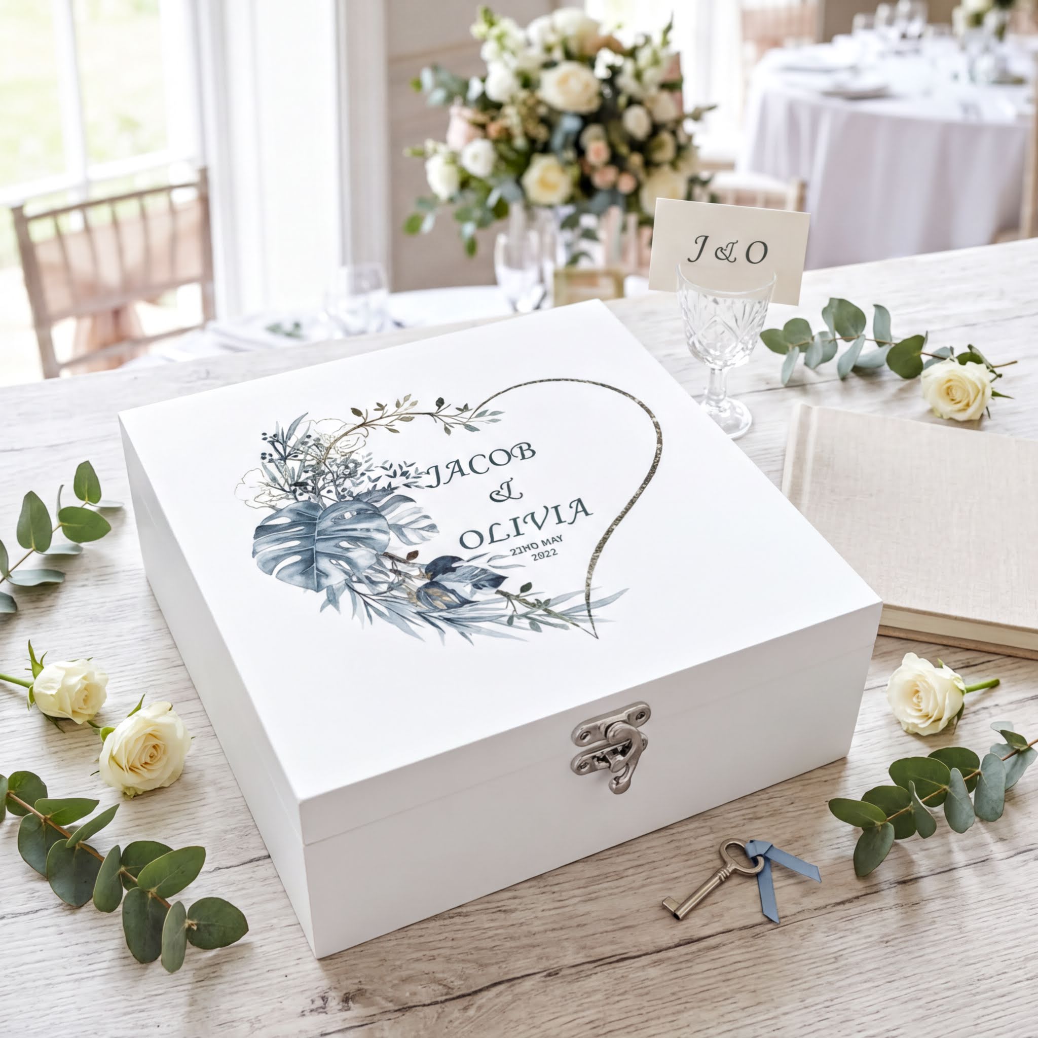 A white wooden box with a floral design and the names " Jacob & Olivia" printed on it, surrounded by white roses and greenery.