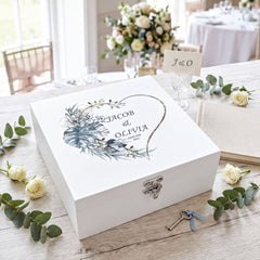 A white wooden box with a floral design and the names " Jacob & Olivia" printed on it, surrounded by white roses and greenery.