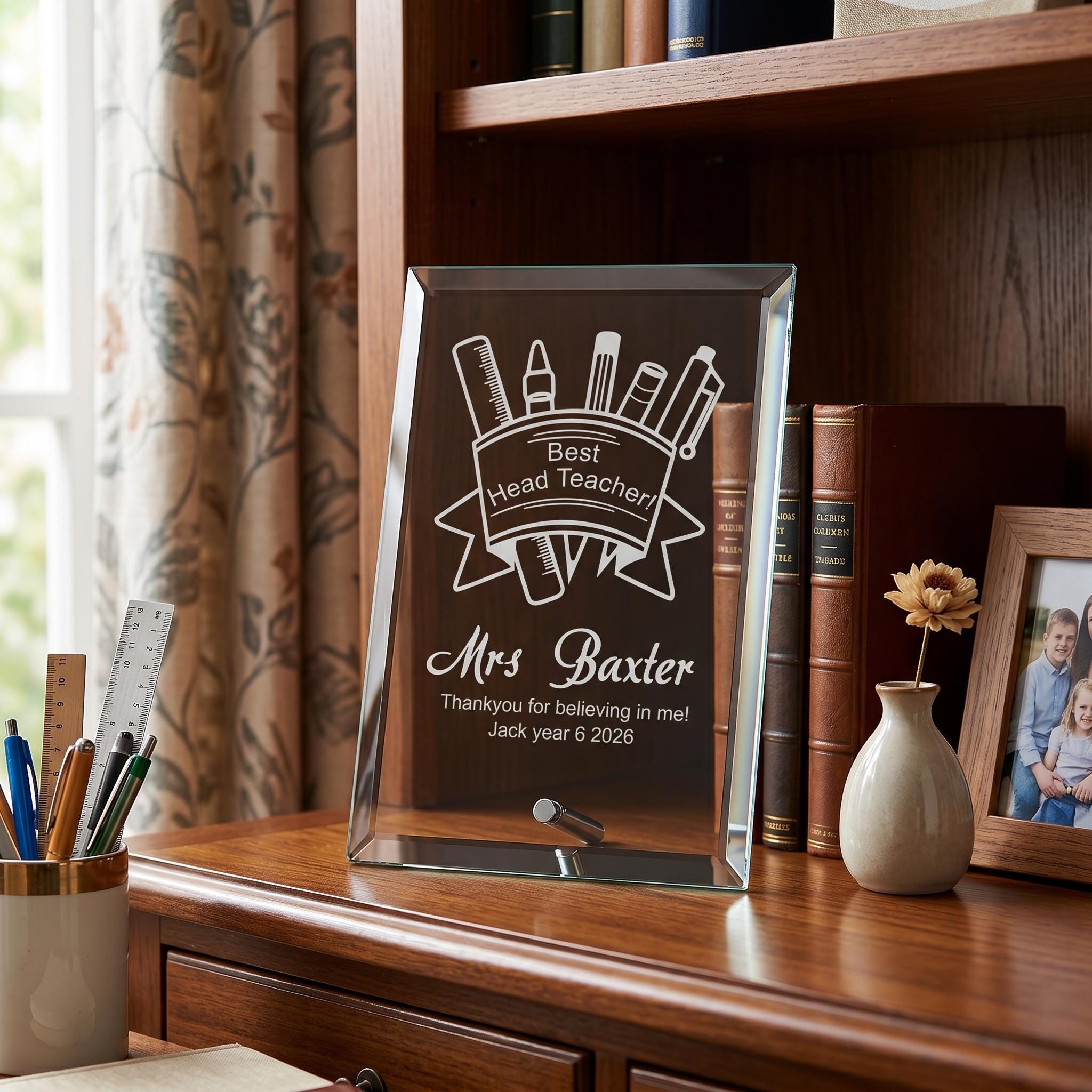 A glass plaque on a wooden desk, displaying a "Best Heading Teacher" message and the name "Mrs Baxter". The plaque is surrounded by various items, including a vase with a flower, books, and a ruler.