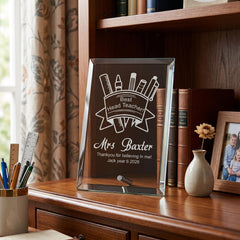 A glass plaque on a wooden desk, displaying a "Best Heading Teacher" message and the name "Mrs Baxter". The plaque is surrounded by various items, including a vase with a flower, books, and a ruler.