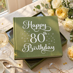 A green book titled "Happy 80th Birthday" sits on a table surrounded by flowers and a glass of wine.