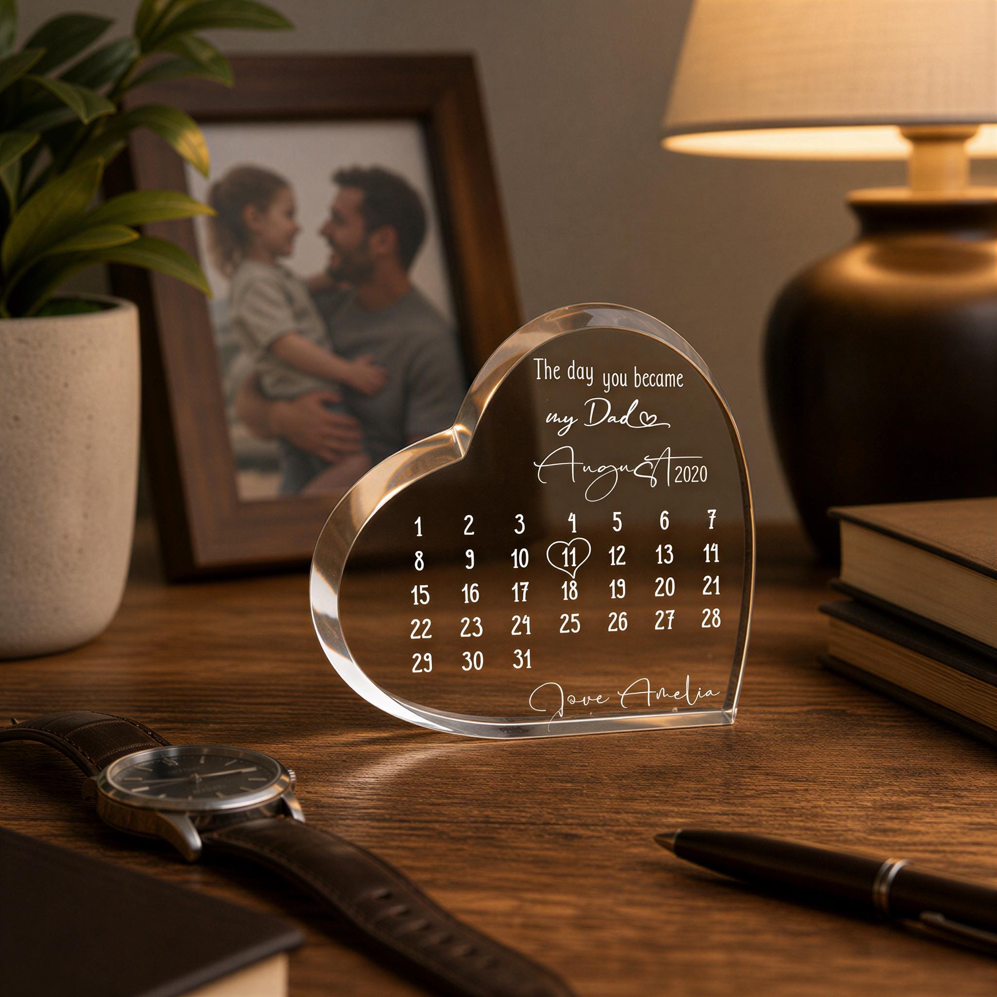 a heart-shaped glass desk calendar with a photo of a man and a child on it, placed on a wooden desk with a lamp and a book.