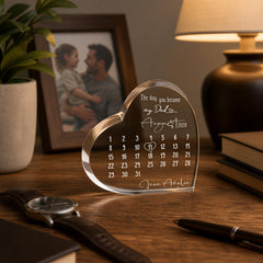 a heart-shaped glass desk calendar with a photo of a man and a child on it, placed on a wooden desk with a lamp and a book.