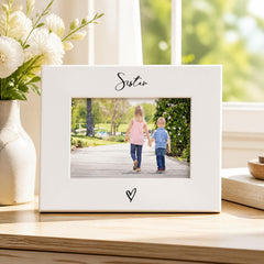 A white picture frame with a photo of two children walking together, placed on a wooden surface next to a vase of white flowers.
