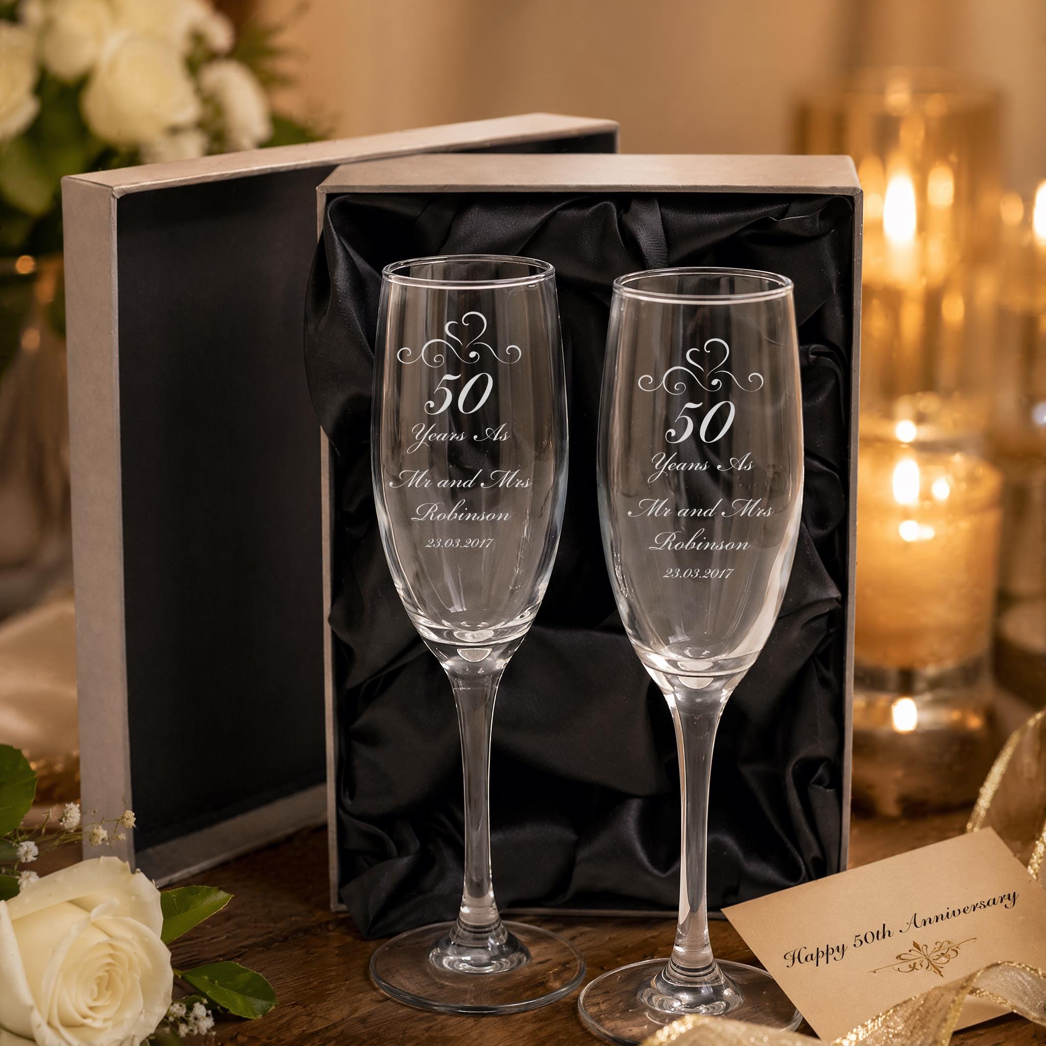 a pair of elegant champagne glasses, each engraved with a message celebrating a 50th anniversary. The glasses are placed on a table, with a card and some flowers visible in the background.
