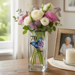 A vase filled with a bouquet of white and pink flowers, including roses and lavender, sits on a wooden table. The vase has a butterfly design and the number "70" printed on it. In the background, there is a framed photo of an elderly woman.