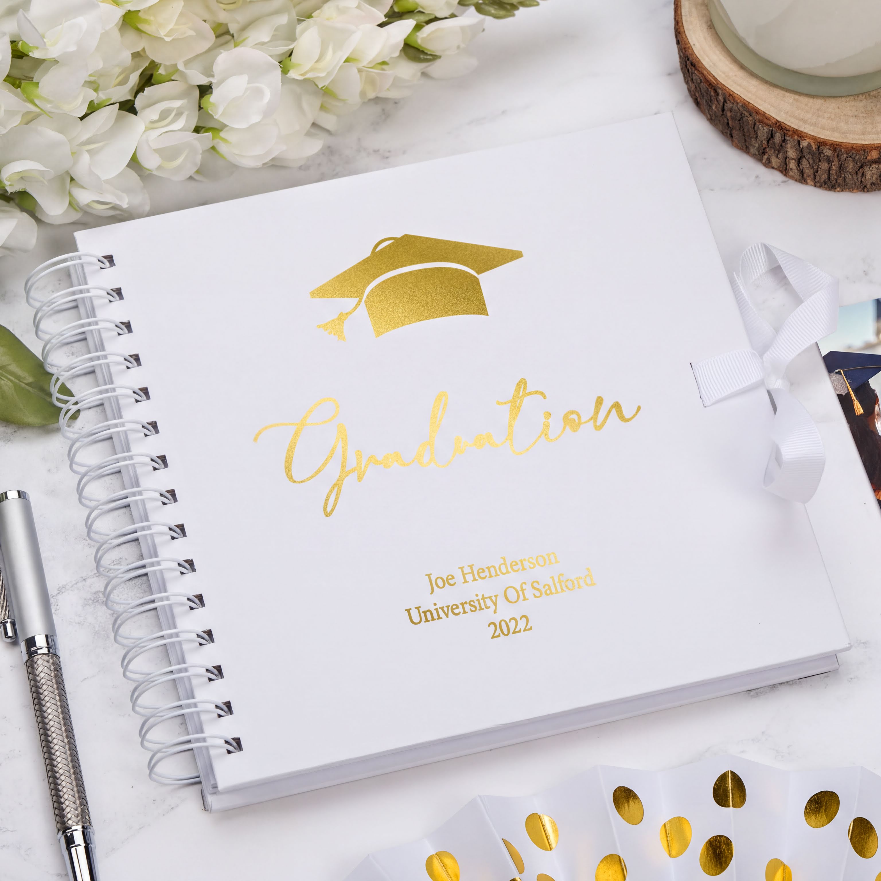 a graduation cap and a graduation cap-themed notebook with the word "Graduation" written on it, placed on a marble surface.