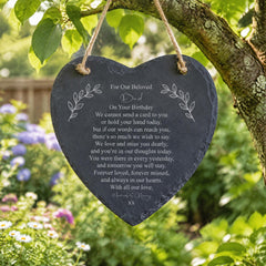A heart-shaped slate hanging from a tree branch with a heartfelt message written on it.
