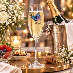 a champagne glass with a personalized message, a bottle of champagne, and a silver bucket with a cork, all set on a table with flowers and other decorative items.