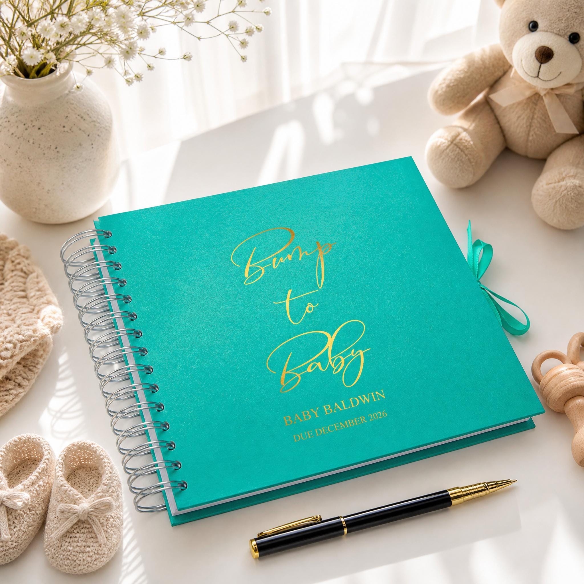A turquoise-colored spiral-bound book titled "Bumps to Baby" sits on a white surface, accompanied by a teddy bear, a vase with flowers, and a pen.