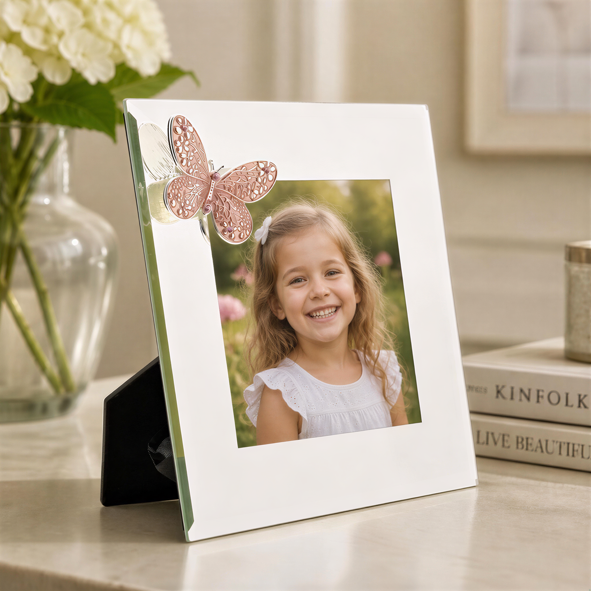 A young girl with a bright smile is the focus of a white picture frame with a butterfly decoration, placed on a table with a vase of white flowers in the background.