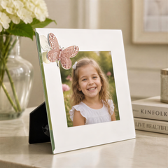 A young girl with a bright smile is the focus of a white picture frame with a butterfly decoration, placed on a table with a vase of white flowers in the background.