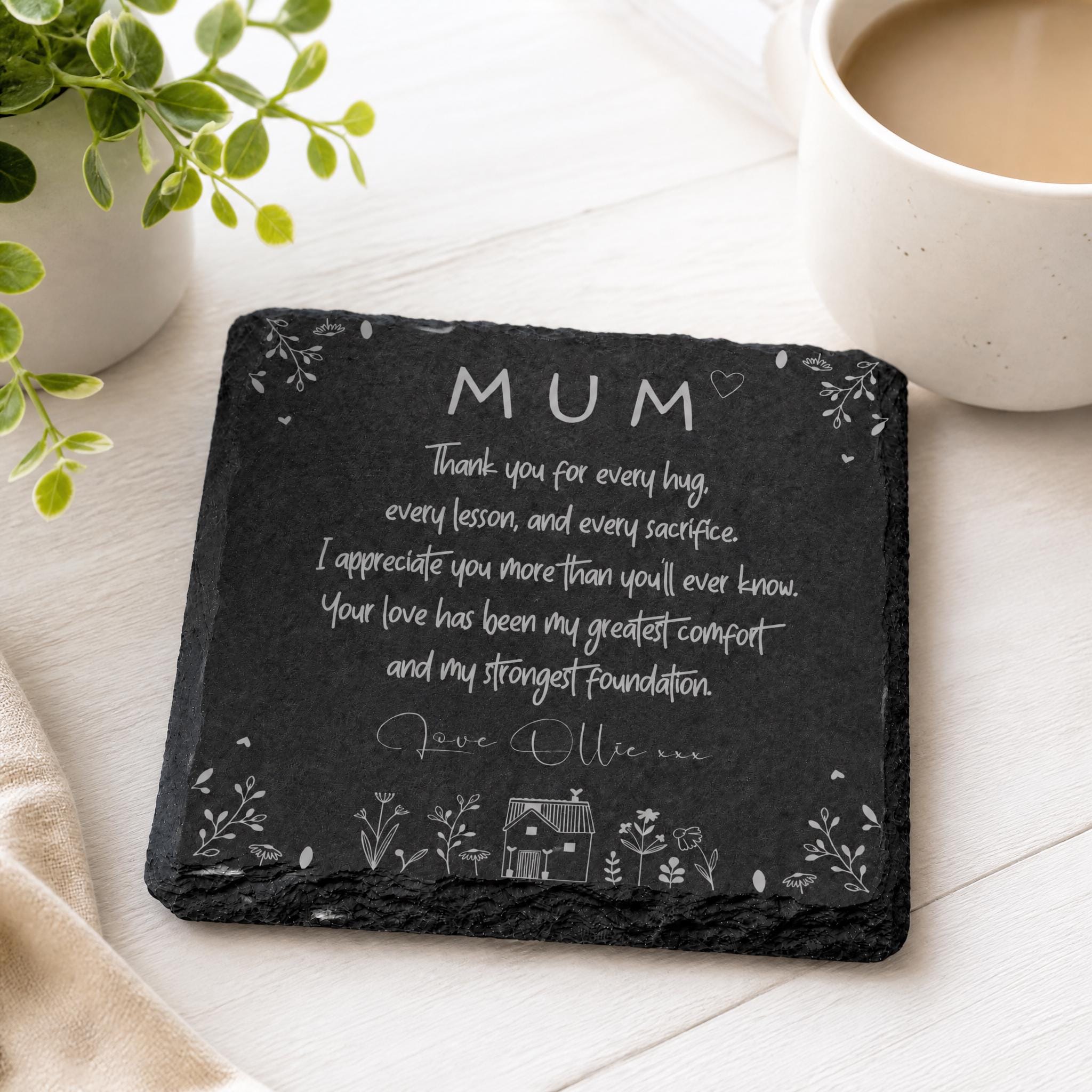 A black slate coaster with a heartfelt message written on it, placed on a white surface next to a cup of coffee and a potted plant.