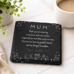 A black slate coaster with a heartfelt message written on it, placed on a white surface next to a cup of coffee and a potted plant.