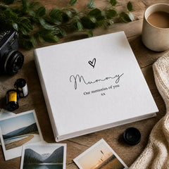 A white photo album with the text "Mummy Our memories of you" and a heart symbol sits on a wooden surface, surrounded by various items such as a camera, a mug, and some photographs.