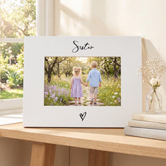 A white picture frame with a photograph of two children, a boy and a girl, standing in a garden surrounded by flowers. The word "Sister" is written above the photograph.