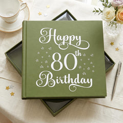 A green book titled "Happy 80th Birthday" sits on a table, surrounded by a teacup, a vase of flowers, and scattered gold stars.