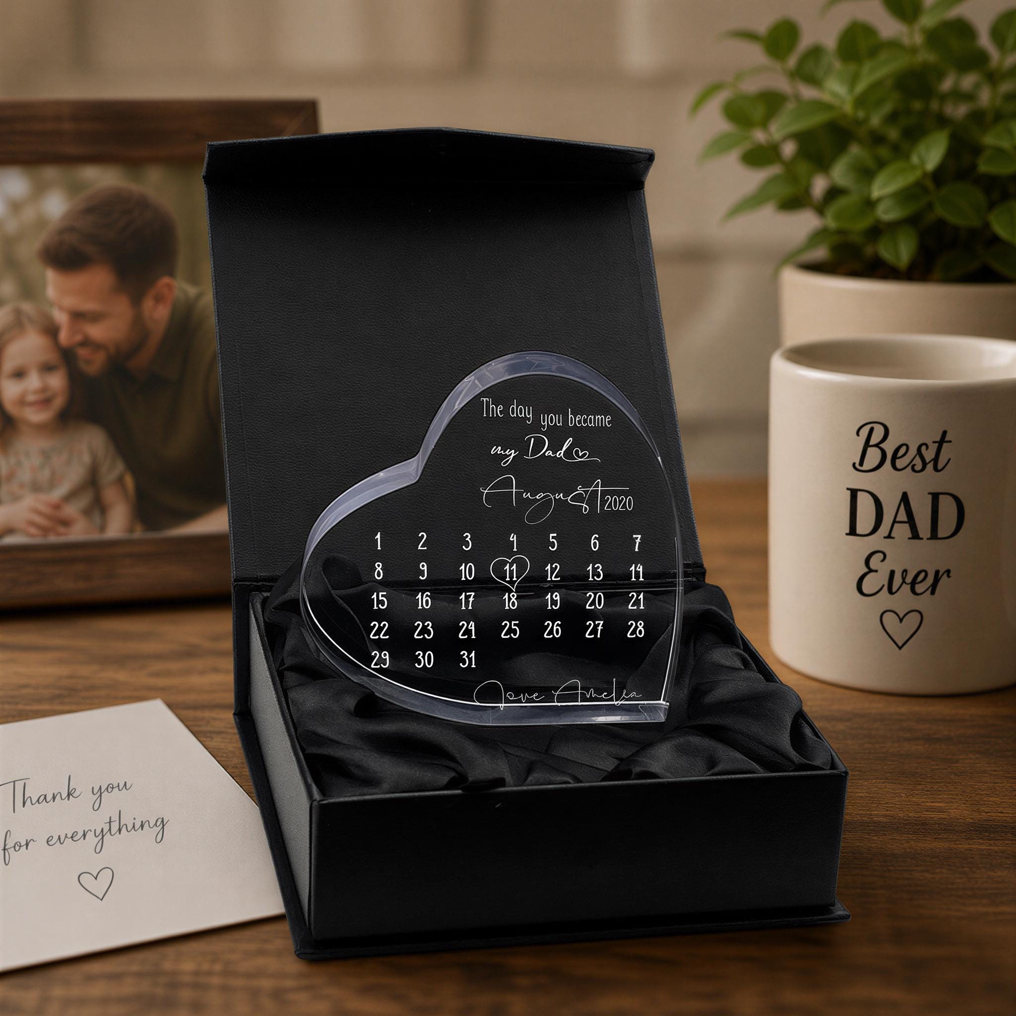 a heart-shaped calendar with a personal message, placed on a wooden surface next to a mug with a "Best Dad Ever" inscription. There is also a photo frame and a plant in the background.
