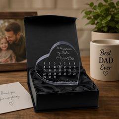 a heart-shaped calendar with a personal message, placed on a wooden surface next to a mug with a "Best Dad Ever" inscription. There is also a photo frame and a plant in the background.