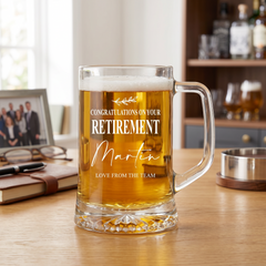 A personalized glass mug filled with beer, placed on a wooden table, with a framed photo and other items in the background.