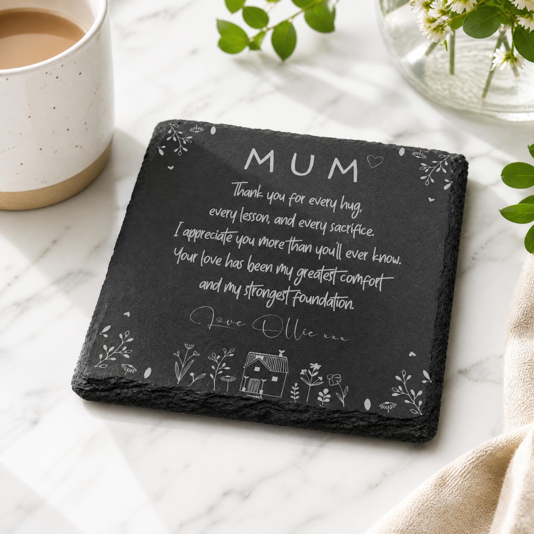 A black slate coaster with a heartfelt message written on it, placed on a marble surface next to a white mug and a vase of flowers.