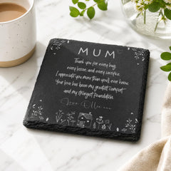 A black slate coaster with a heartfelt message written on it, placed on a marble surface next to a white mug and a vase of flowers.
