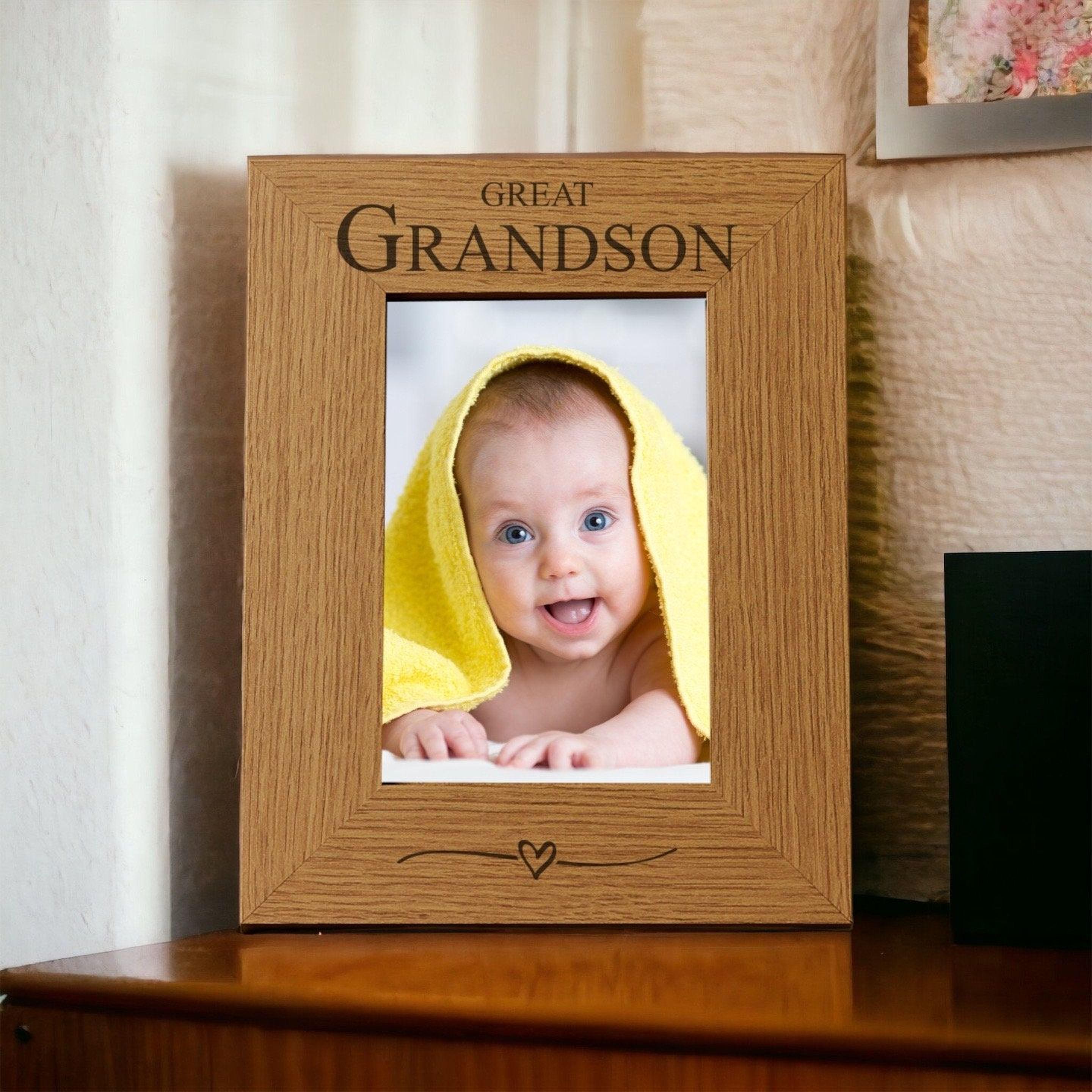 A wooden picture frame with a photo of a smiling baby wrapped in a yellow towel inside it. The frame has the text "GREAT GRANDSON" written on it.