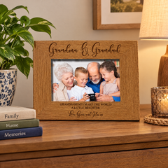 A wooden picture frame with a family photo of a man, woman, and two children, sitting on a wooden surface. The frame has a message that reads "Grandma & Grandpa" and "Grandparents make the world a little brighter".
