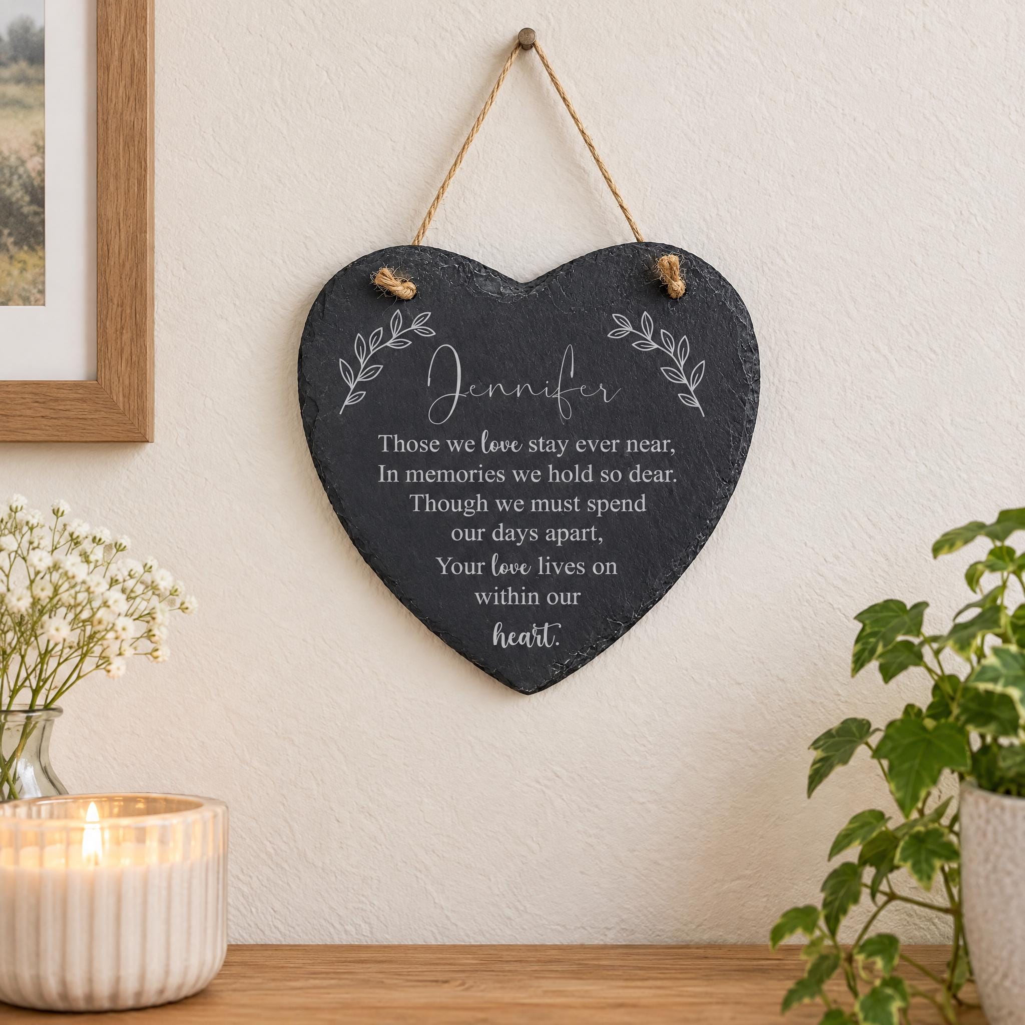 a heart-shaped slate hanging on a wall, with a candle in a white vase to the left and a potted plant to the right.