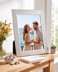 A man and a woman are standing together in a white picture frame, smiling and looking at each other, with a scenic view of the ocean and a balcony in the background.