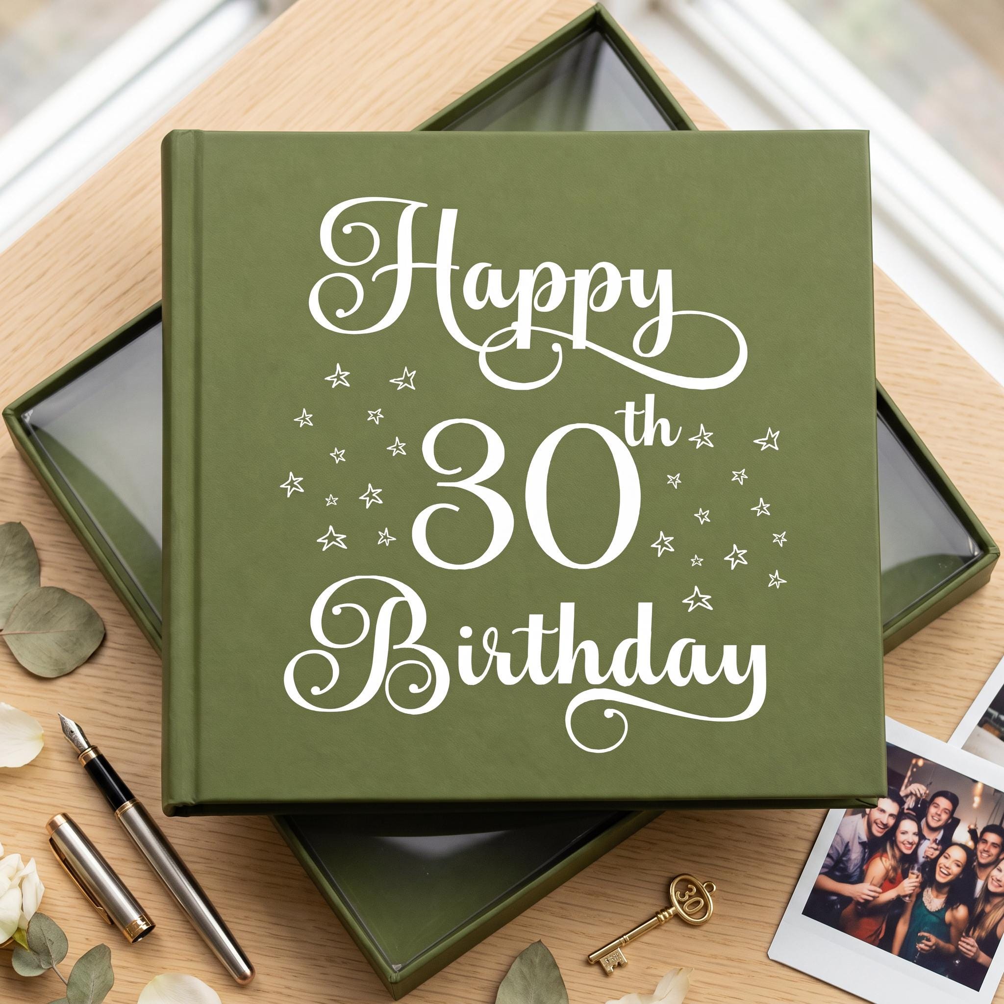 A green photo album with the text "Happy 30th Birthday" printed on it, placed on a wooden surface alongside some flowers and a key.