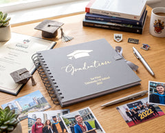 a graduation cap, a graduation certificate, a graduation book, and various graduation-related items such as photographs and pens, all arranged on a wooden surface.