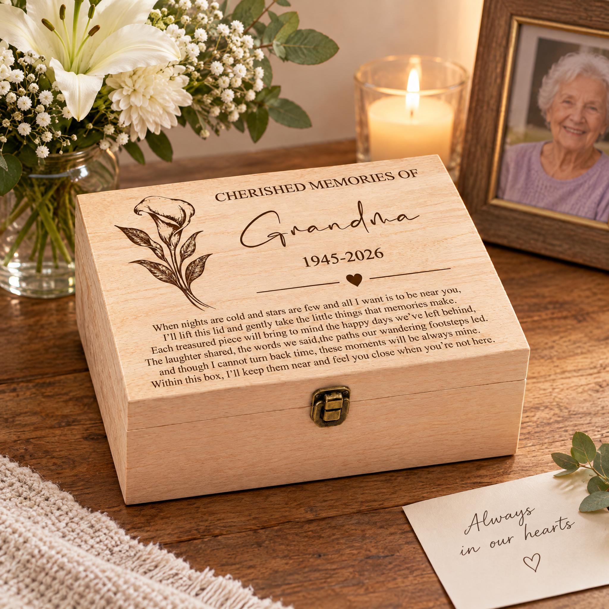 a wooden box with a heartfelt message engraved on it, placed on a wooden surface. The box is accompanied by a note that reads "Always in our hearts". In the background, there is a vase filled with white flowers, and a framed photograph of an elderly woman.