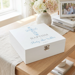 A white wooden box with a cross design and the text "Baptism" is placed on a wooden table, accompanied by a vase of flowers and a framed photo.