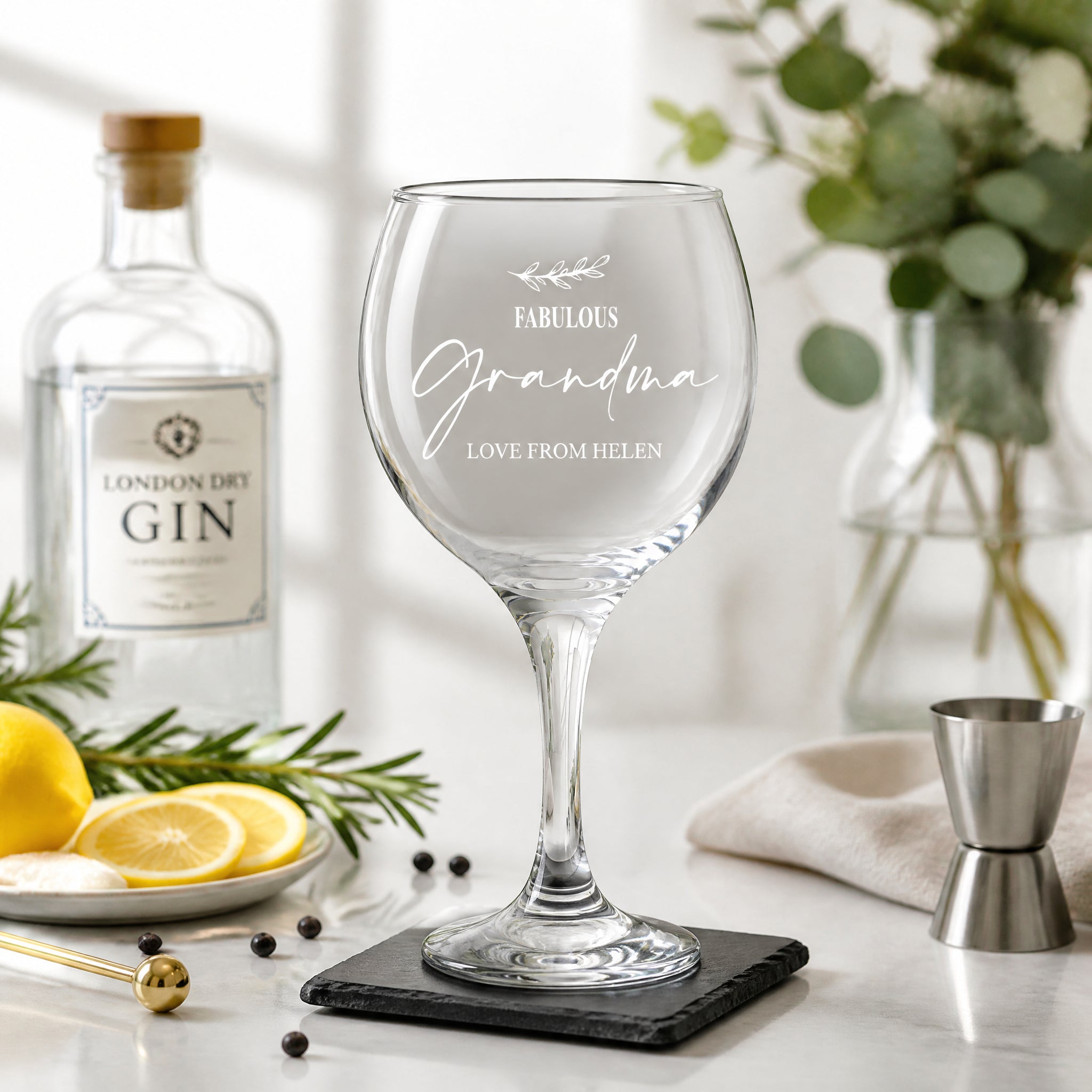 a personalized glass of wine with a message engraved on it, placed on a table with various items such as a bottle of gin, a lemon, and a vase of flowers.