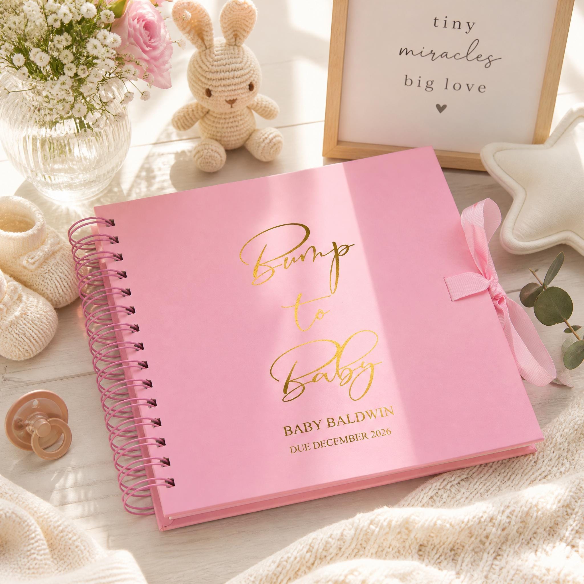 A pink baby book titled "Bump to Baby" by Baby Baldwin, with a pink ribbon and a stuffed bunny rabbit, sits on a wooden surface alongside a framed sign that says "tiny miracles big love".