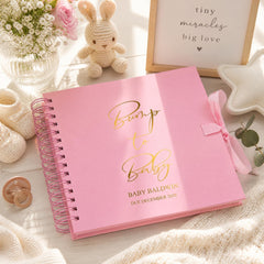 A pink baby book titled "Bump to Baby" by Baby Baldwin, with a pink ribbon and a stuffed bunny rabbit, sits on a wooden surface alongside a framed sign that says "tiny miracles big love".