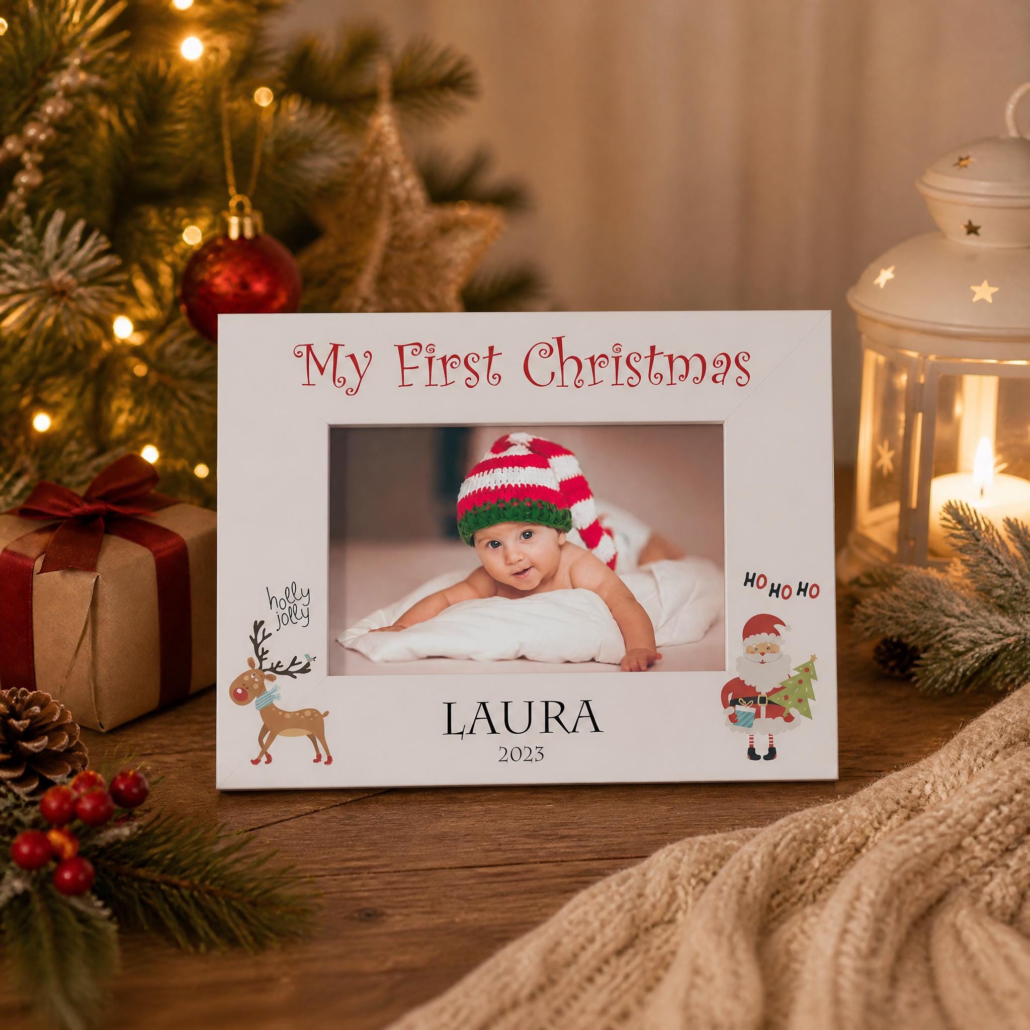 A white picture frame with a photo of a baby wearing a red and white striped hat, surrounded by holiday decorations such as a Christmas tree, ornaments, and a lantern.