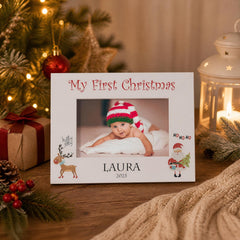 A white picture frame with a photo of a baby wearing a red and white striped hat, surrounded by holiday decorations such as a Christmas tree, ornaments, and a lantern.