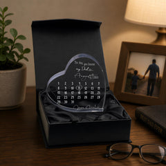 a heart-shaped photo frame containing a calendar, placed on a wooden surface next to a potted plant and a framed photograph.