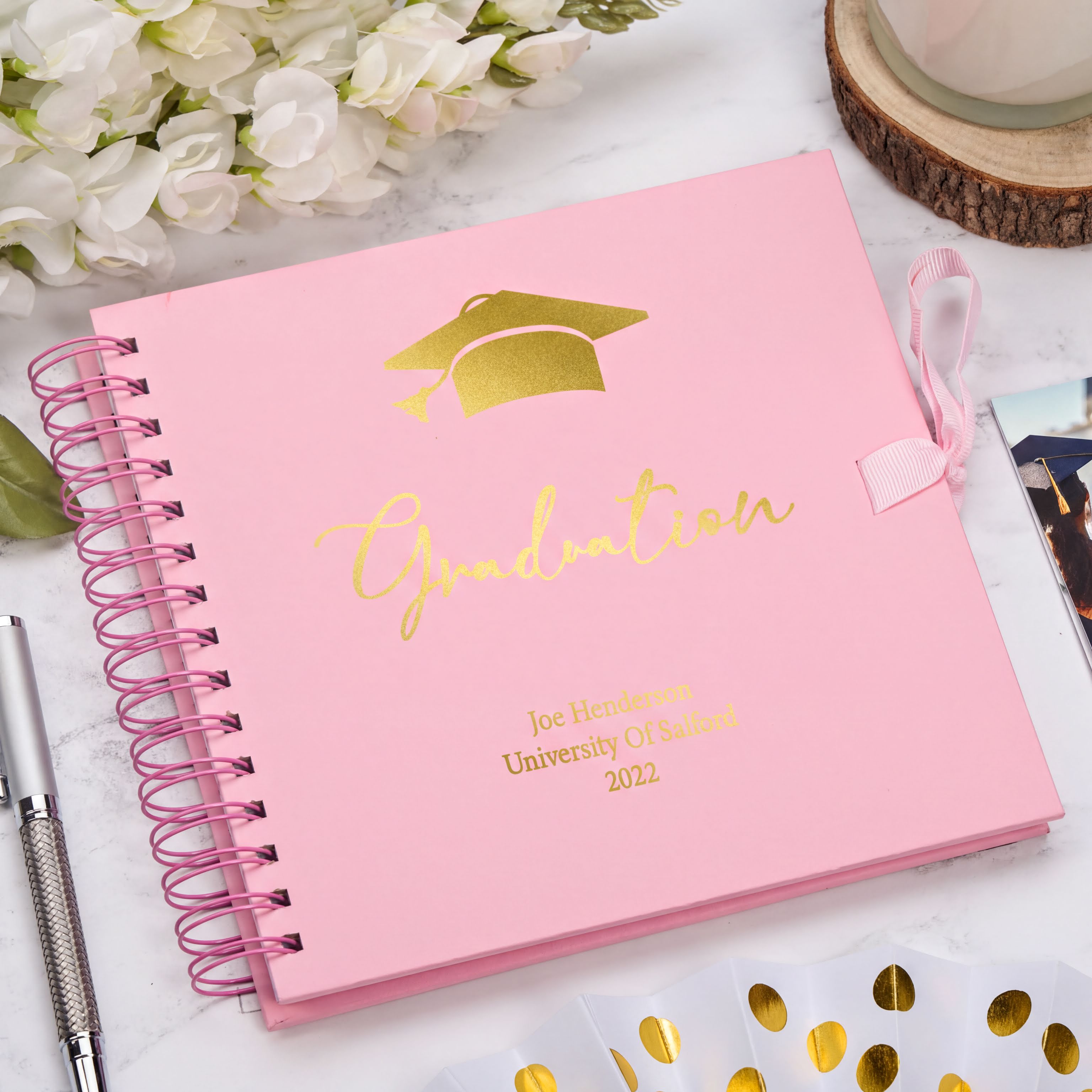 A pink graduation yearbook with gold text and a graduation cap sits on a marble surface, surrounded by white flowers, a pen, and a wooden coaster.