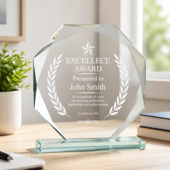 A clear glass award trophy with a star and laurel wreath design, placed on a wooden desk. The trophy is engraved with the text "EXCELLENCE AWARD Presented to John Smith In recognition of your outstanding dedication, leadership and achievement 1st September 2026".