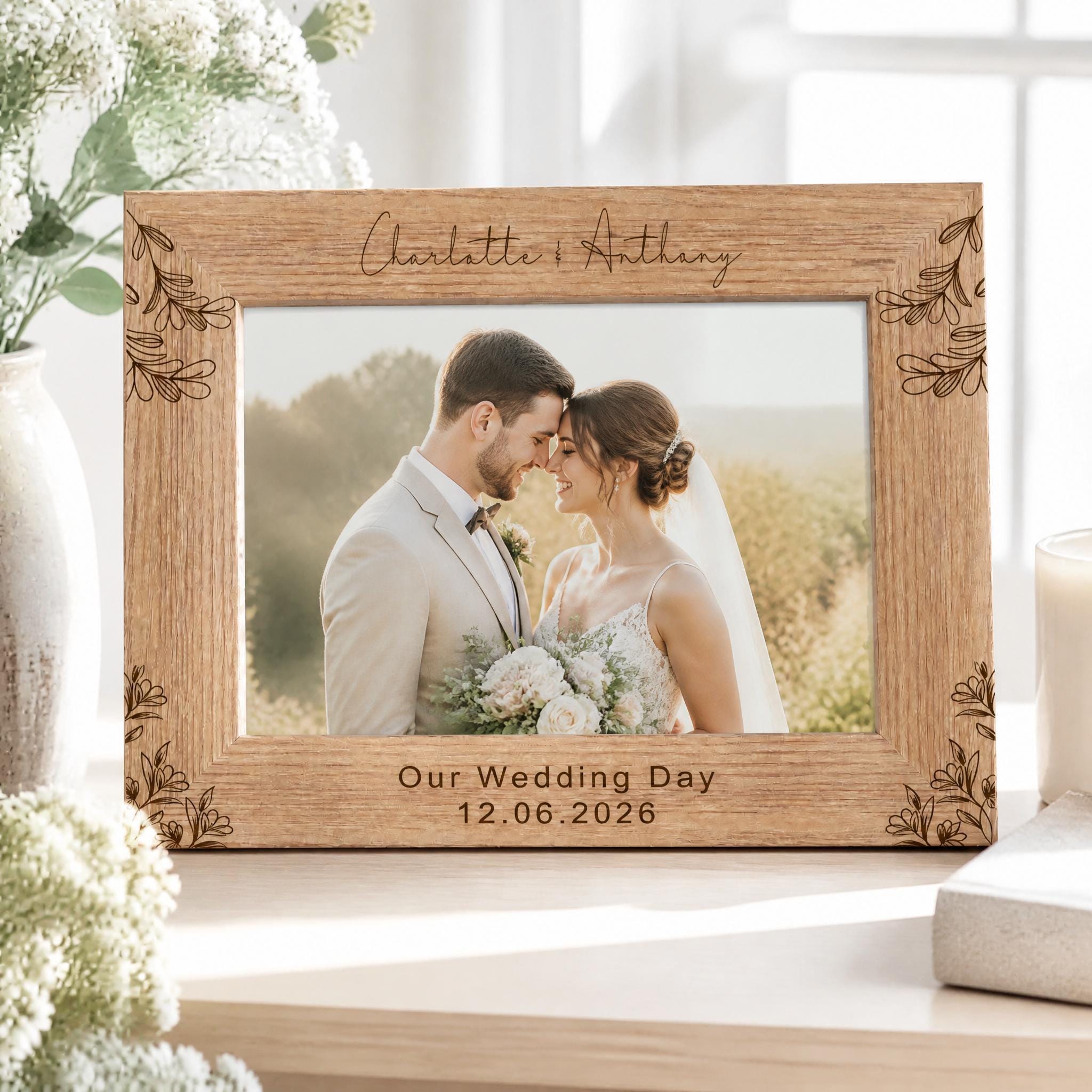 A wooden picture frame with a photo of a couple, presumably a bride and groom, inside it. The frame has a floral design and the names "Charlotte & Anthony" written on it. The photo appears to be a wedding picture, as indicated by the date "Our Wedding Day 12.06.2026" printed below the photo.