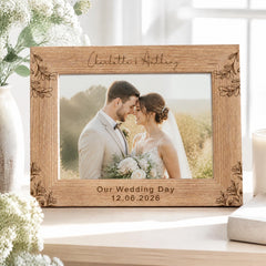 A wooden picture frame with a photo of a couple, presumably a bride and groom, inside it. The frame has a floral design and the names "Charlotte & Anthony" written on it. The photo appears to be a wedding picture, as indicated by the date "Our Wedding Day 12.06.2026" printed below the photo.