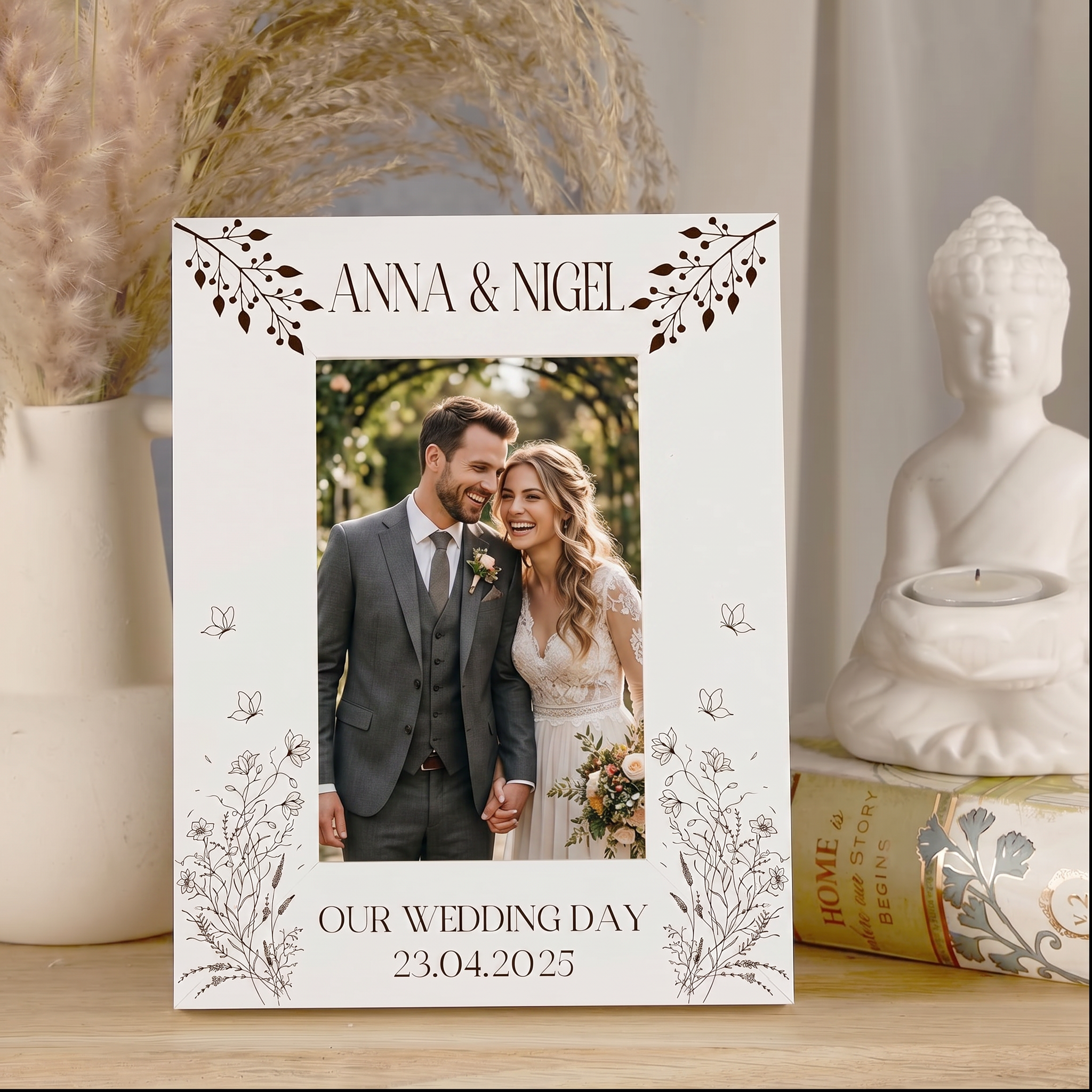 A white wedding picture frame with a couple's photograph inside, surrounded by decorative elements such as a Buddha statue and a vase with flowers.